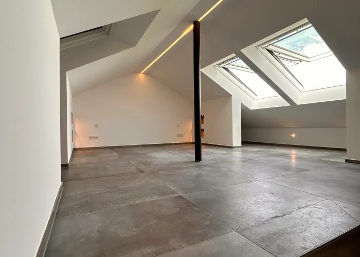 Leerer Dachraum mit Betonboden, weißen Wänden, schwarzer Stütze in der Mitte, eingelassener LED-Lichtleiste an der Decke und zwei großen Dachfenstern